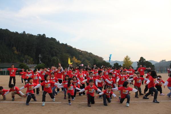 運動場で、赤いティーシャツを着た子ども達が両手を広げ片足を一歩前に出し、ポースを取っており、後方にいる人々も両手を上げて楽しんでいる様子の写真